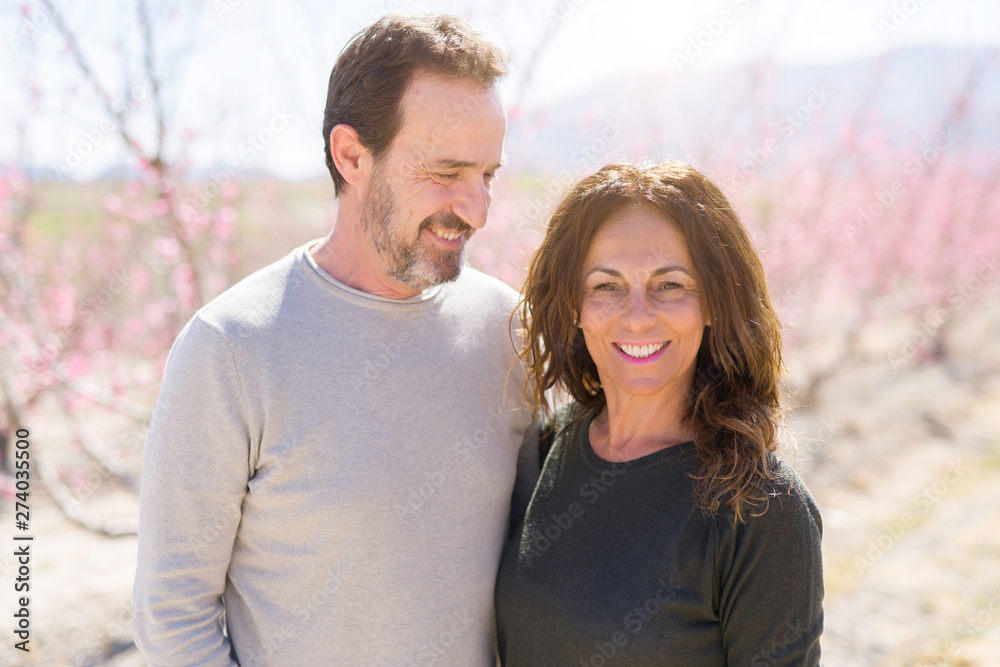 Beautiful middle age senior couple smiling in love at romantic garden of peach trees with pink petals on a sunny day of spring