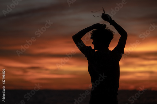 Medium long shot silhouette freedom young man traveler wearing sunglass standing on ocean island mountain cliff at summer sunset with blurred sunset twilight sky and ocean background.