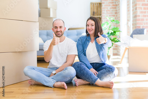 Young couple sitting on the floor arround cardboard boxes moving to a new house doing happy thumbs up gesture with hand. Approving expression looking at the camera with showing success.