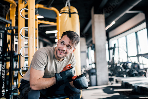 Smiling millennial man in boxing gloves on a break during workout at the gym.