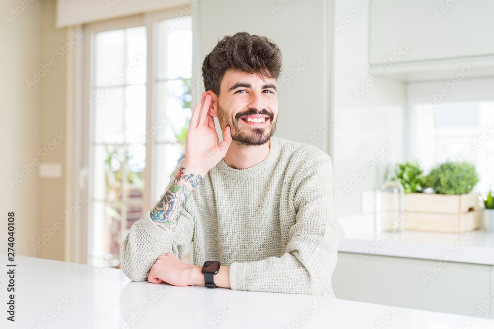 Young man wearing casual sweater sitting on white table smiling with hand over ear listening an hearing to rumor or gossip. Deafness concept.