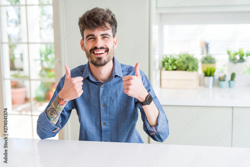 Young man wearing casual shirt sitting on white table success sign doing positive gesture with hand, thumbs up smiling and happy. Looking at the camera with cheerful expression, winner gesture. Young man wearing casual shirt sitting on white table success sign doing positive gesture with hand, thumbs up smiling and happy. Looking at the camera with cheerful expression, winner gesture.
