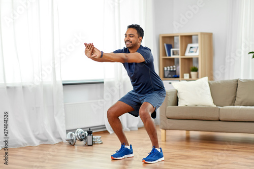 Fototapeta Naklejka Na Ścianę i Meble -  sport and healthy lifestyle concept - smiling indian man with fitness tracker exercising and doing squats at home