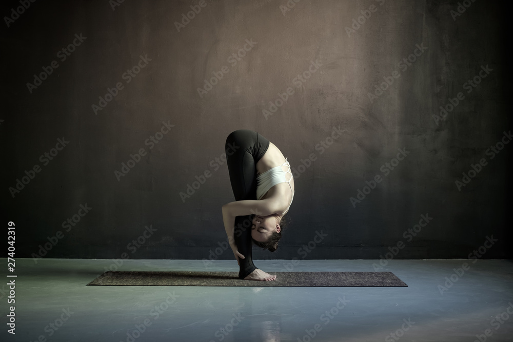Young european woman practicing yoga, doing head to knees, uttanasana exercise, Standing forward bend pose. Indoor full length, black wall in yoga studio