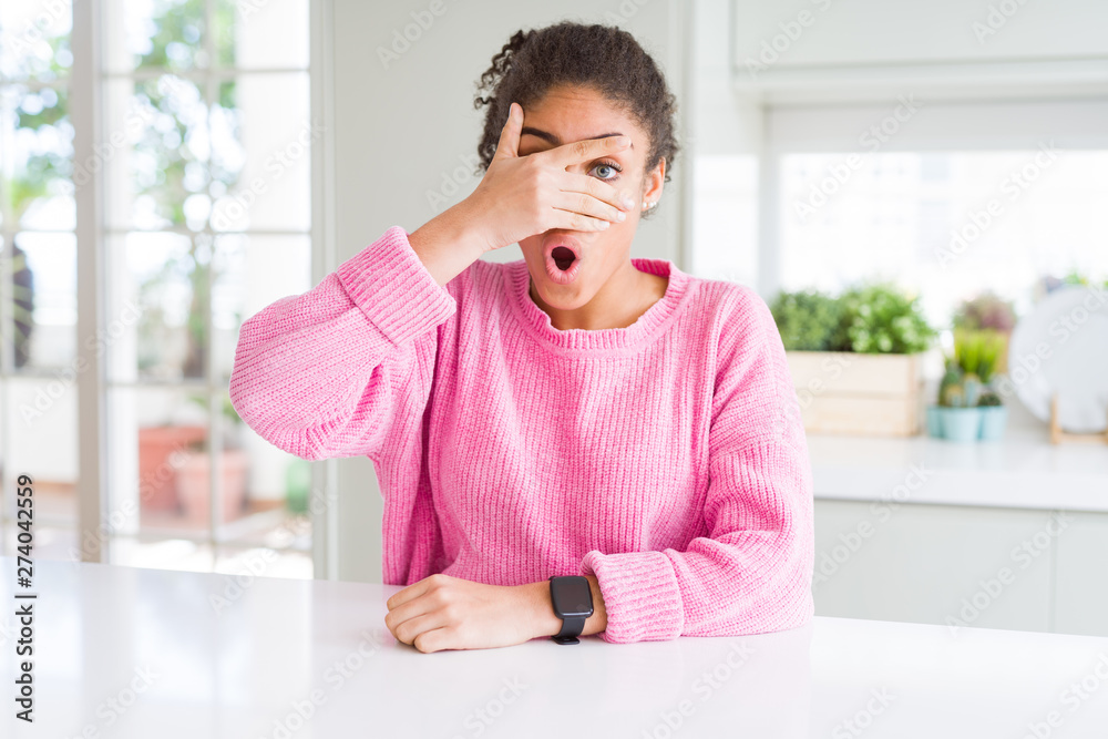 Beautiful african american woman with afro hair wearing casual pink sweater peeking in shock covering face and eyes with hand, looking through fingers with embarrassed expression.