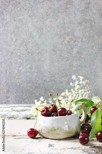 Fresh cherry berries in ceramic mug, elderflowers, jug of cream on old wooden kitchen table with gray wall at background. Copy space