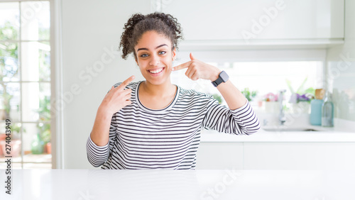 Beautiful african american woman with afro hair wearing casual striped sweater smiling cheerful showing and pointing with fingers teeth and mouth. Dental health concept.