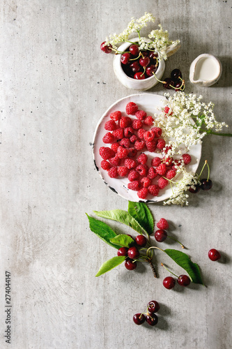 Fresh cherry and raspberry berries in ceramic mug and plate, elderflowers, jug of cream over gray texture background. Flat lay, space