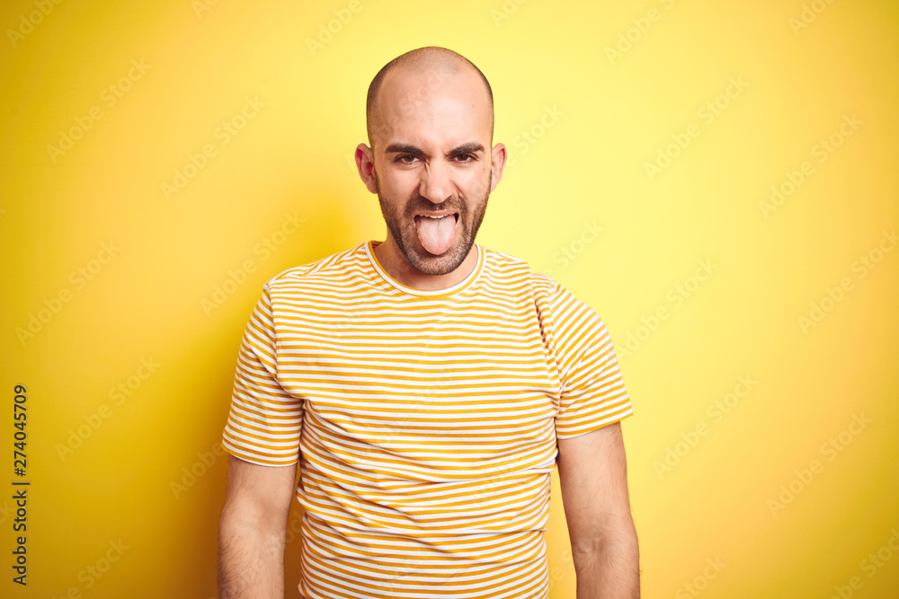 Young bald man with beard wearing casual striped t-shirt over yellow isolated background sticking tongue out happy with funny expression. Emotion concept. Young bald man with beard wearing casual striped t-shirt over yellow isolated background sticking tongue out happy with funny expression. Emotion concept.