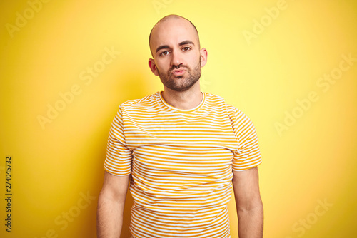 Young bald man with beard wearing casual striped t-shirt over yellow isolated background looking at the camera blowing a kiss on air being lovely and sexy. Love expression.