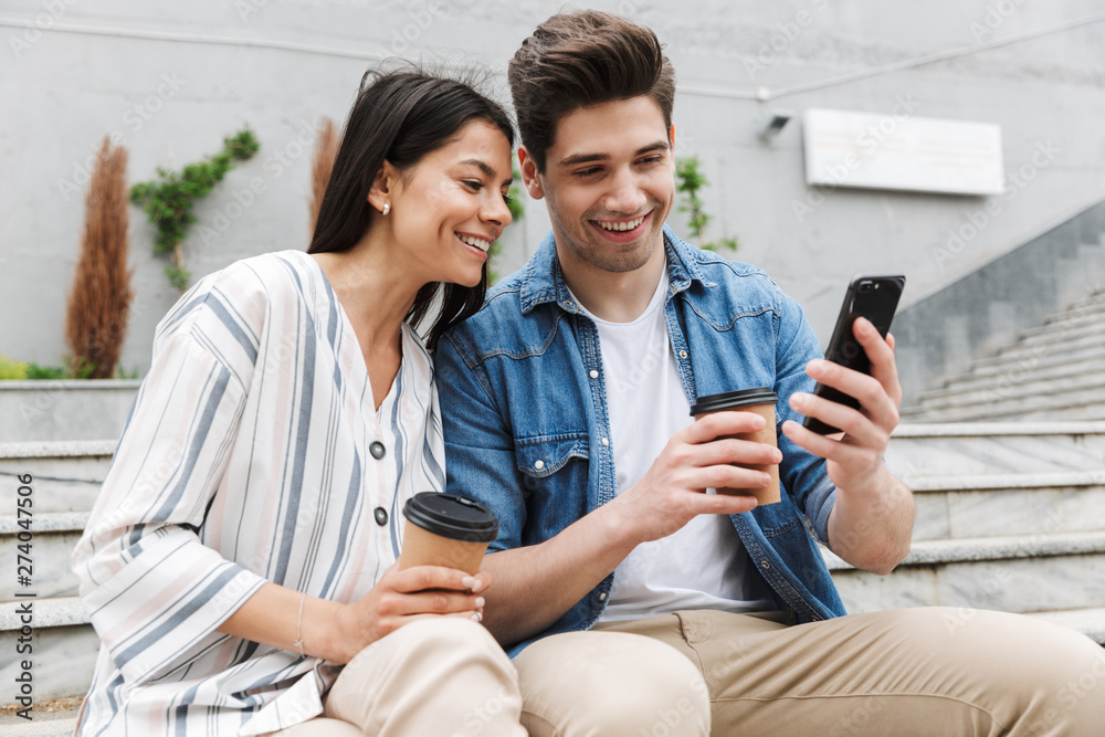 Image of cheery couple drinking takeaway coffee and using smartphone on city stairs outdoors Image of cheery couple drinking takeaway coffee and using smartphone on city stairs outdoors