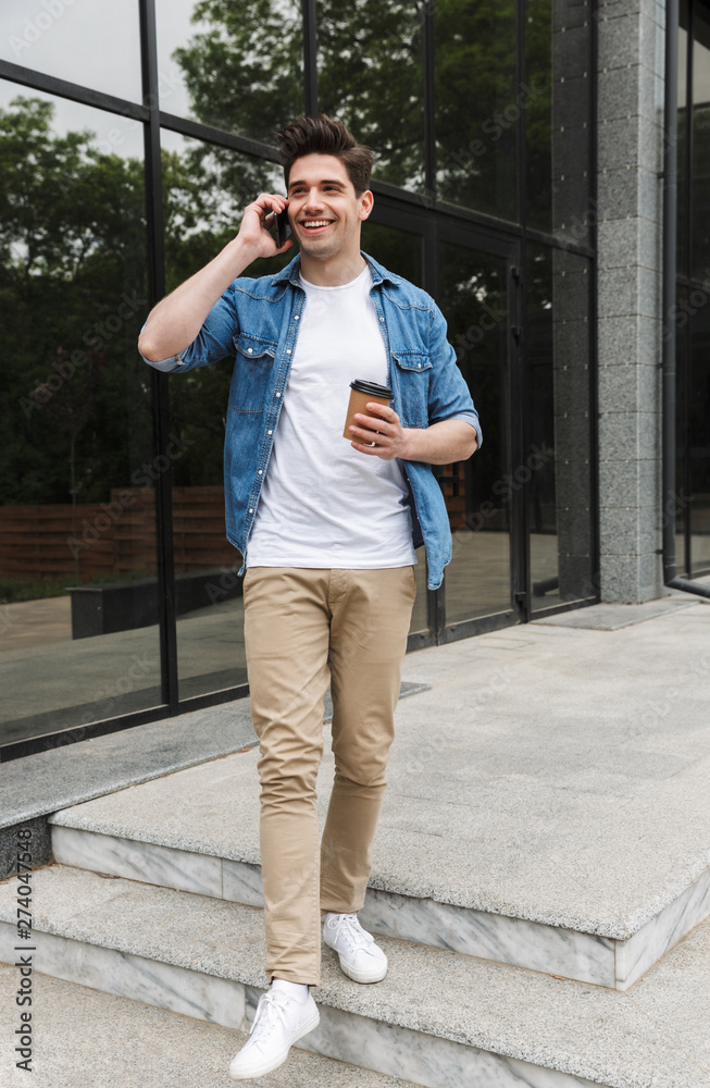 Image of cheery young man drinking takeaway coffee and talking on cellphone while standing over building