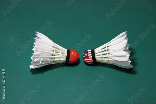 Two used shuttlecocks on green floor of Badminton court with both head each other. One head painted with China flag and one head painted with the Malaysia flag.