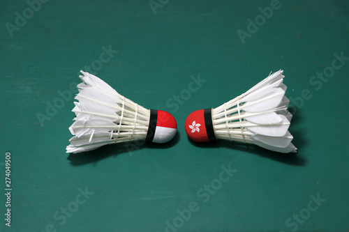 Two used shuttlecocks on green floor of Badminton court with both head each other. One head painted with Indonesia flag and one head painted with the Hong Kong flag.
