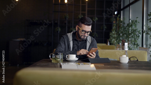 handsome hipster man is typing on screen of smartphone sitting in restaurant in daytime, tablet in front