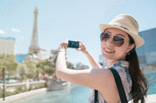 cheerful asian female tourist in sunglasses taking photo of eiffel tower in paris with smartphone. young smiling girl backpacker in straw hat looking in camera. happy woman travel in europe in summer