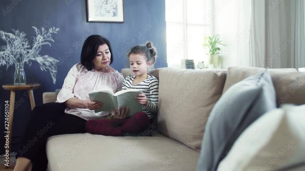A small girl with grandmother sitting on sofa at home, reading a book. A small girl with grandmother sitting on sofa at home, reading a book.