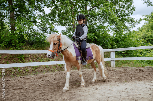 Fototapet Caucasian girl with helmet and protective vest on riding cute white and brown pony horse