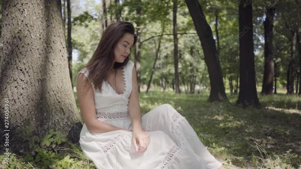 Portrait of cute young girl with long brunette hair wearing a long white summer fashion dress sitting under a tree in the park. Leisure of a pretty woman looking at the camera outdoors. Slow motion. Portrait of cute young girl with long brunette hair wearing a long white summer fashion dress sitting under a tree in the park. Leisure of a pretty woman looking at the camera outdoors. Slow motion.