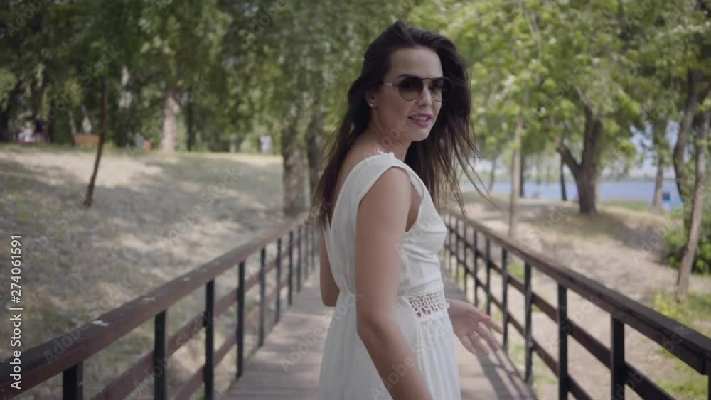 Portrait glamorous brunette young girl wearing sunglasses and long white summer fashion dress walking along wooden bridge. Pretty woman looking at the camera outdoors. Follow me. Gesture. Slow motion. Portrait glamorous brunette young girl wearing sunglasses and long white summer fashion dress walking along wooden bridge. Pretty woman looking at the camera outdoors. Follow me. Gesture. Slow motion.