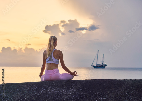 Young blonde slender woman doing yoga meditation on the cliff by the sea at sunrise       