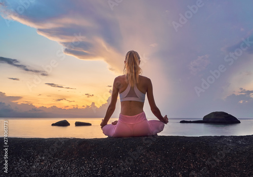 Young blonde slender woman doing yoga meditation on the cliff by the sea at sunrise 