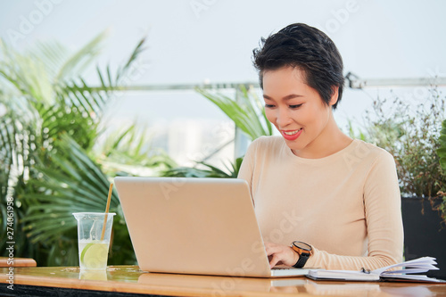 Asian young businesswoman sitting at the table typing on laptop computer and smiling while working in cafe