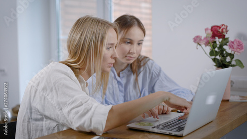Portrait of a two young women sitting in kitchen and surfing web on laptop, having fun on internet, best friends.