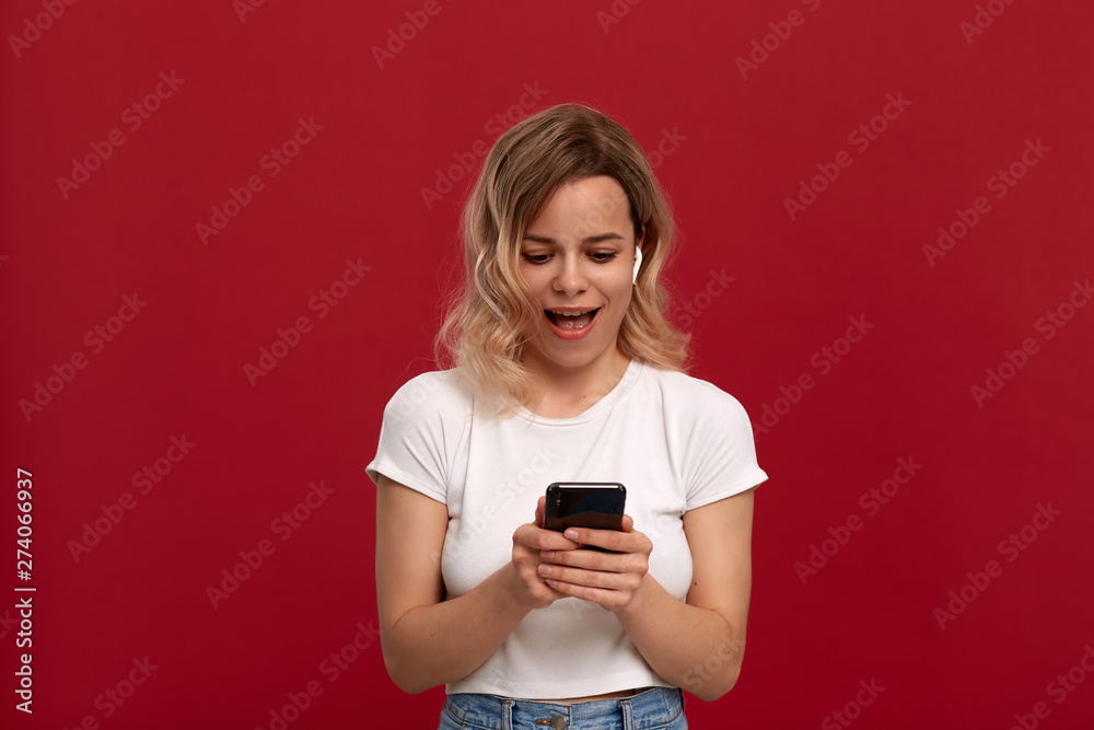 Portrait of a girl with curly blond hair in a white t-shirt on a red background. Happy model looks at the screen of a mobile phone. Portrait of a girl with curly blond hair in a white t-shirt on a red background. Happy model looks at the screen of a mobile phone.