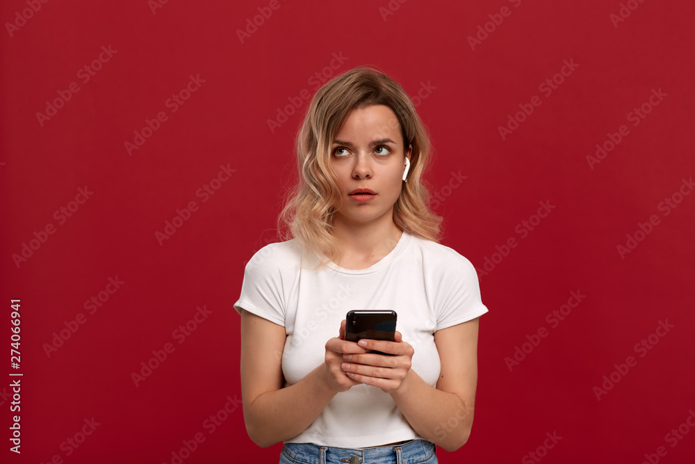 Portrait of a girl with curly blond hair in a white t-shirt on a red background. Surprised model in wireless headset shows screen of a mobile phone. Portrait of a girl with curly blond hair in a white t-shirt on a red background. Surprised model in wireless headset shows screen of a mobile phone.