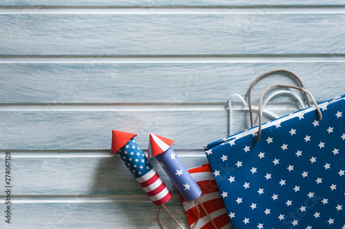 Fototapeta Naklejka Na Ścianę i Meble -  4th July holiday shopping concept. Shopping bags in national american colors and firework rockets over wooden background