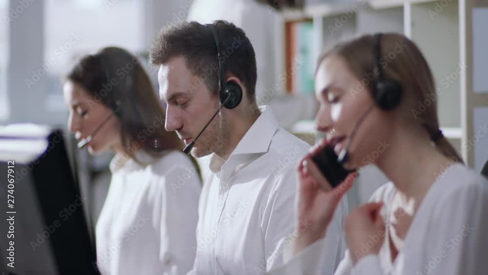 Selective focus on team of professional support call center operators working in the office, female operator gets the call on her smartphone, she answers it and puts off the headset. Profession