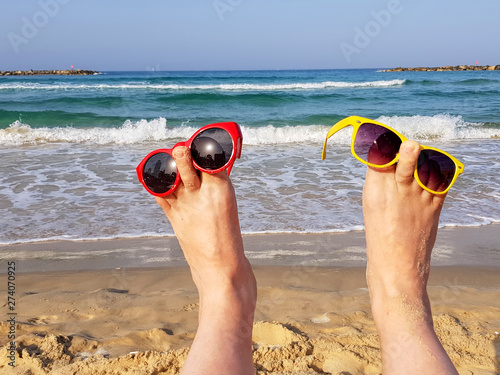 Bare feet raised up with colored sunglasses on the beach. Beautiful sea surf and blue sunny sky in background. Summer vacation. Funny imagination happy character 