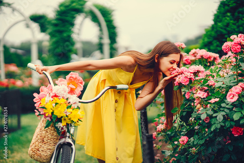 a cute and stylish girl in a yellow dress and long hair rides on a bicycle with a basket of flowers in the sunny summer garden