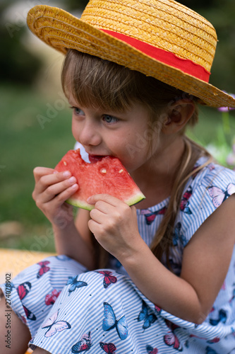 little beautiful girl of 4 years old in a dress and hat is eating a watermelon in the summer in the park at a picnic and smiling