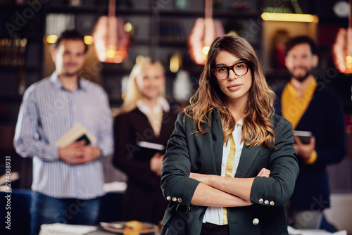 Serious Caucasian businesswoman with long brown hait and dressed smart casual standing in restaurand with crossed arms. In background her successful team posing.