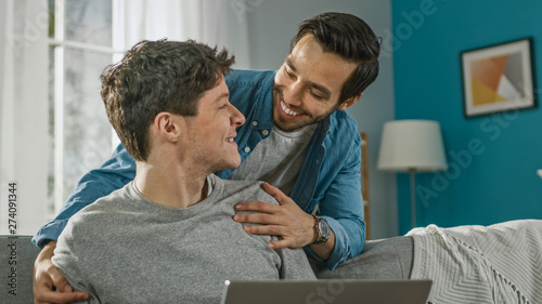Sweet Male Gay Couple Spend Time at Home. Young Man Works on a Laptop, His Partner Comes From Behind and Gently Embraces Him. They Laugh. Room Has Modern Interior.