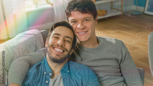 Portrait of Cute Male Queer Couple at Home. They Sit on Sofa and Look at Camera. Partner Embraces His Lover from Behind. They are Happy and Smiling. Room Has Modern Interior with Rainbow Flare Effect