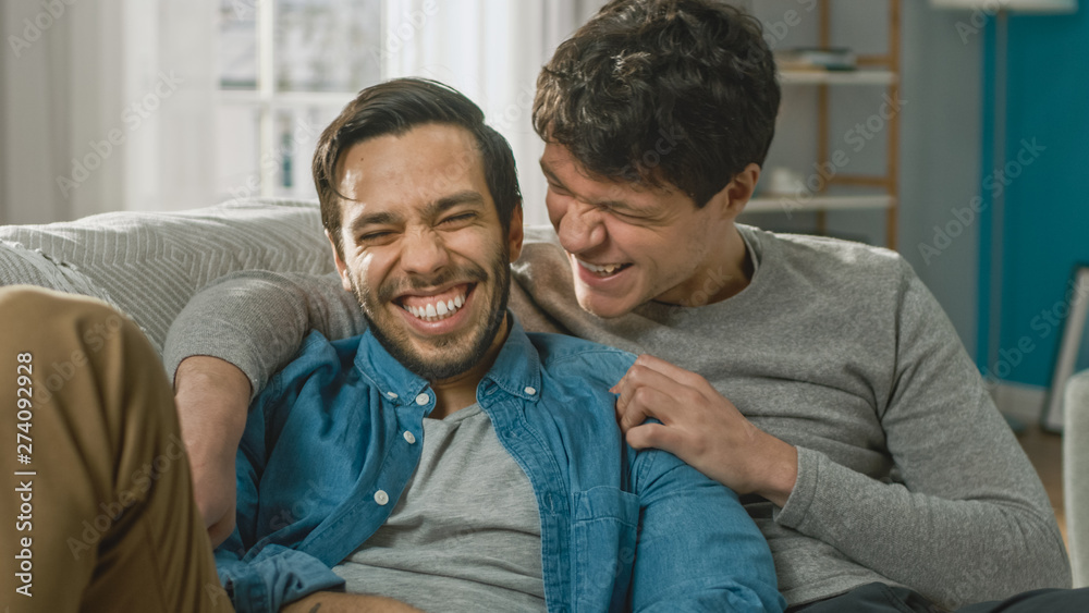 Fototapeta premium Portrait of a Cute Male Queer Couple at Home. They Sit on a Sofa and Joking. Partner Embraces His Lover from Behind. They are Happy and Smiling. Room Has Modern Interior.