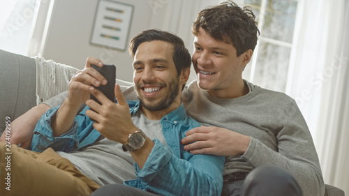 Cute Male Gay Couple Spend Time at Home. They are Lying Down on a Sofa and Use a Smartphone. They Browse Online. Partner's Hand is Around His Lover. They Smile and Laugh. Room Has Modern Interior.