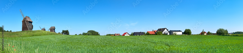 Panoramic image with historical windmill and roofs of rural houses behind wheet field in Spring.