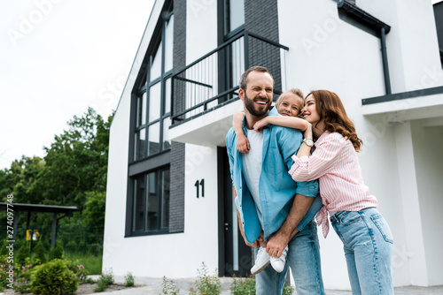 happy bearded man piggybacking daughter near cheerful wife and home photo