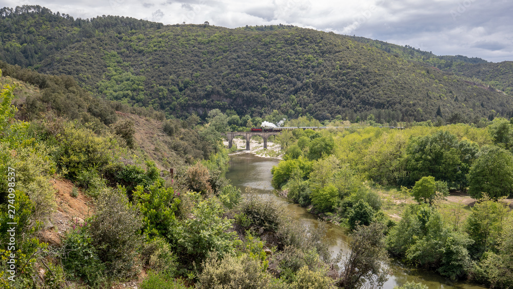 Eine historische Dampflok überquert im Hintergrund auf einer alten Bogenbrücke ein Flusstal in den Cevennen
