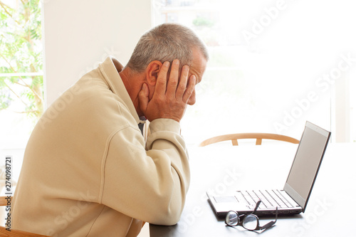 Casual dressed man sitting by a laptop computer is hiding his head in despair. A pair of glasses lying on a pile of papers next to the laptop. - Image