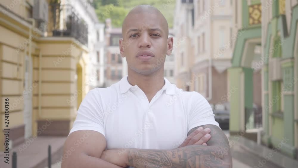 Portrait of handsome confident bald middle eastern man wearing white t-shirt with crossed arms looking in the camera standing on the street in front of old buildings. Portrait of handsome confident bald middle eastern man wearing white t-shirt with crossed arms looking in the camera standing on the street in front of old buildings.