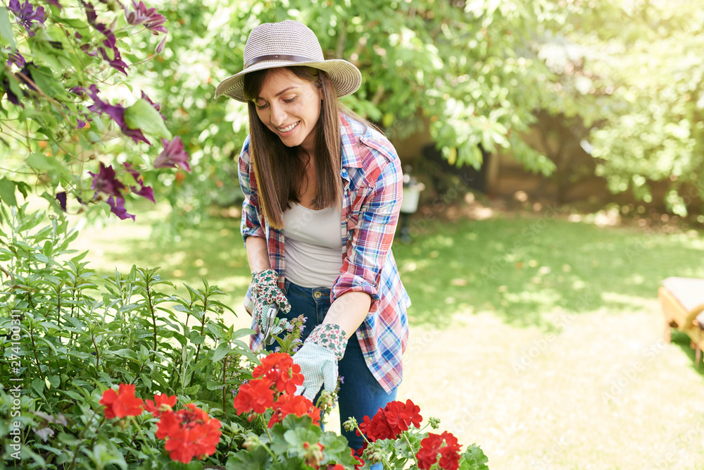 Beautiful smiling Caucasian brunette in working clothes and with hat pruning flowers in her backyard.