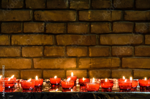 фотографія Small white votive candles lighted and placed in red glass holders on a table covered with aluminium foil in front of a brick wall in a catholic church