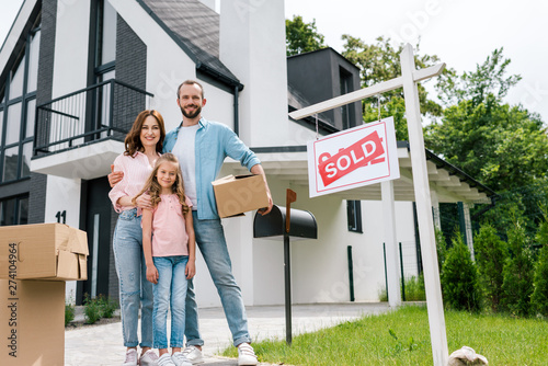happy man holding box and standing with wife and daughter near house and board with sold letters