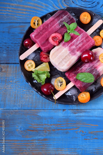 Fruit and berry popsicles on the plate with ice, pieces of kumquat, lime and cherries and mint against the blue background