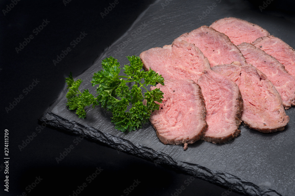 Sliced Grass Fed Juicy Corn Roast Beef garnished with Fresh Curly Parsley on black natural stone background. Sliced Grass Fed Juicy Corn Roast Beef garnished with Fresh Curly Parsley on black natural stone background.